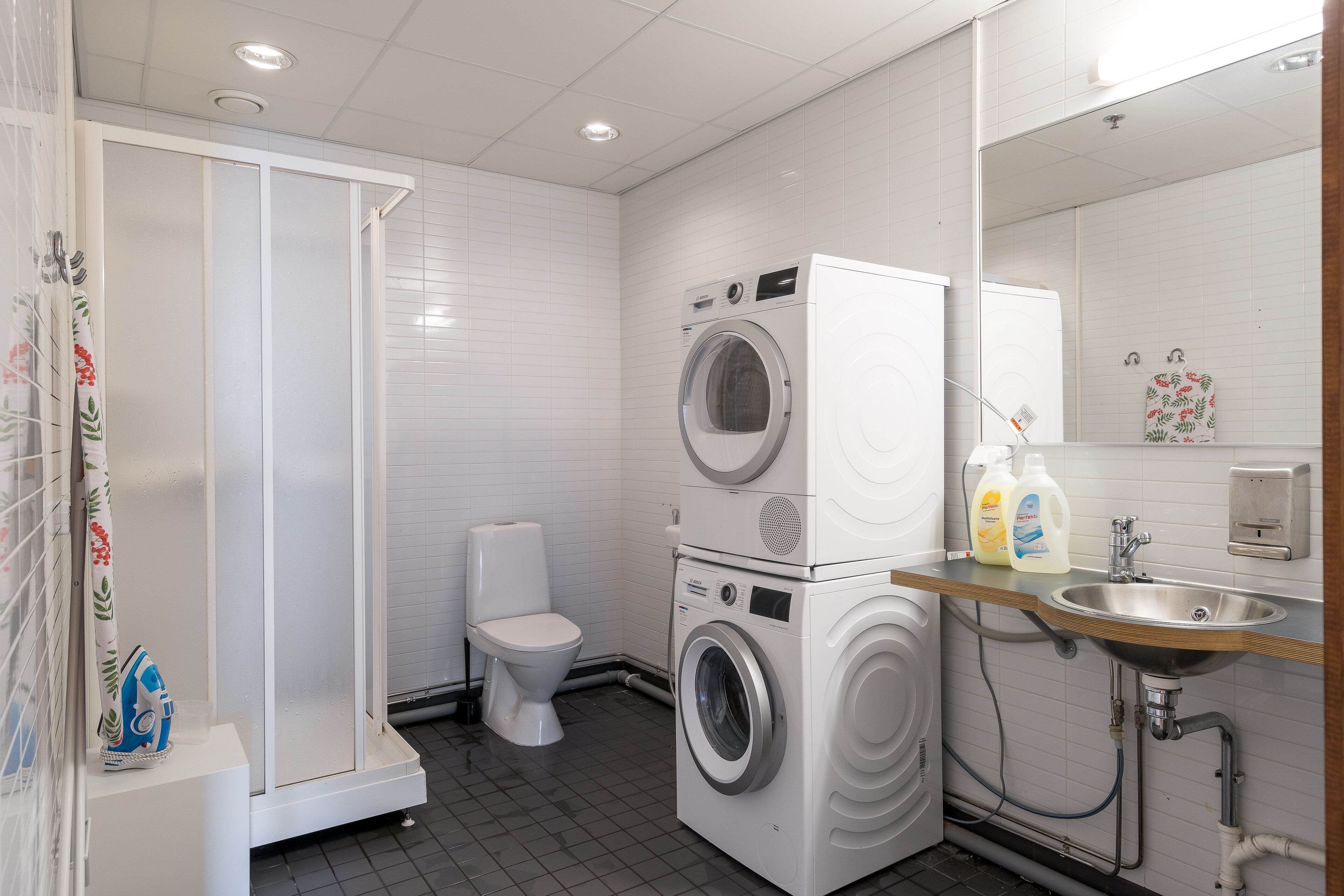 Une petite salle de bain d’hôtel comprenant une douche, des toilettes, un lave-linge et un sèche-linge superposés, ainsi qu’un lavabo avec des distributeurs de savon.
