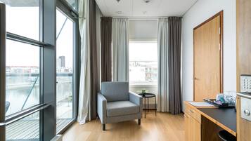A modern hotel room with a large window, a grey armchair, a small round table, and a wooden desk with a computer.