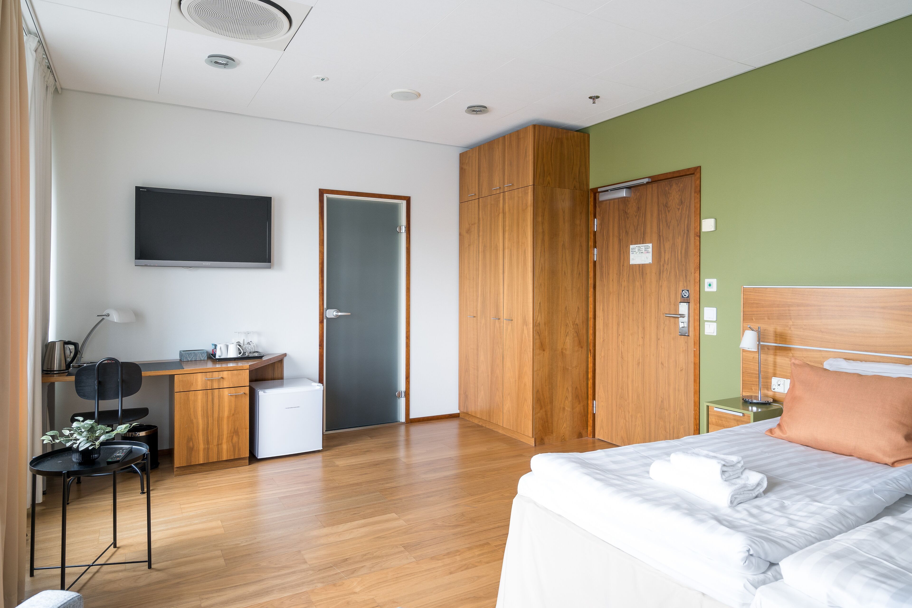 A hotel room with a bed, a desk, a chair, a small kitchenette, and a wardrobe.