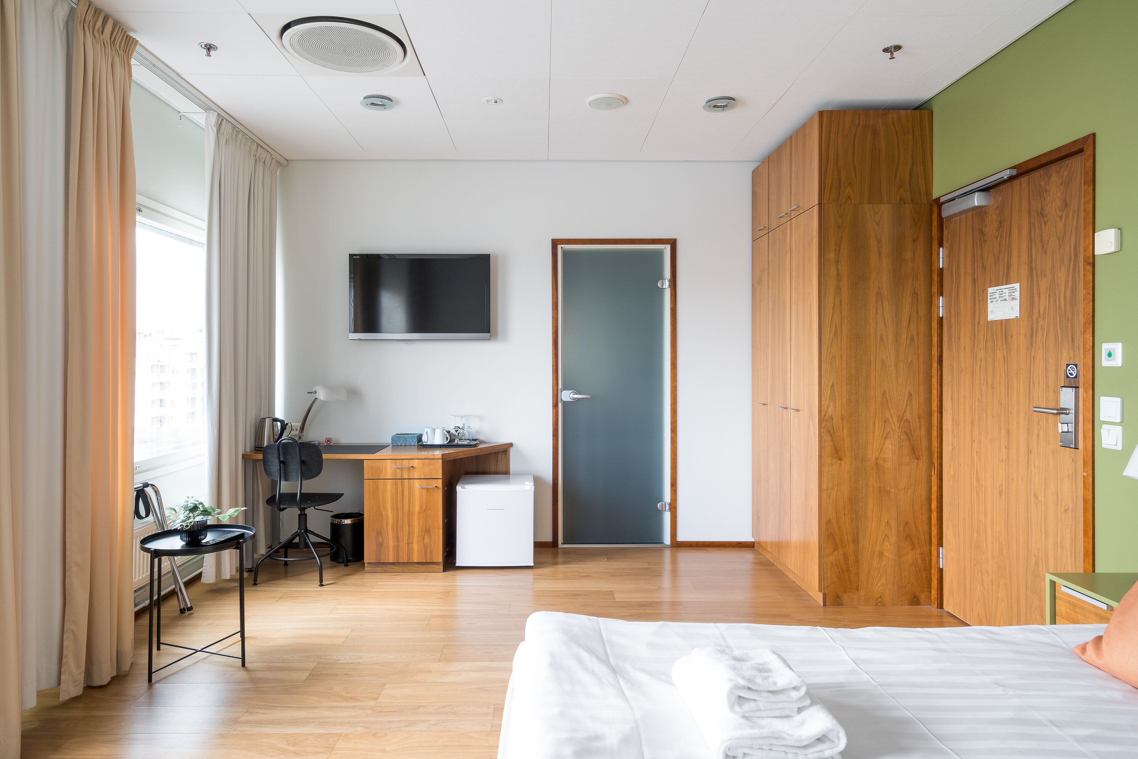 A modern hotel room with a wooden floor, a bed with white linens, a desk with a computer, a chair, and a small refrigerator.