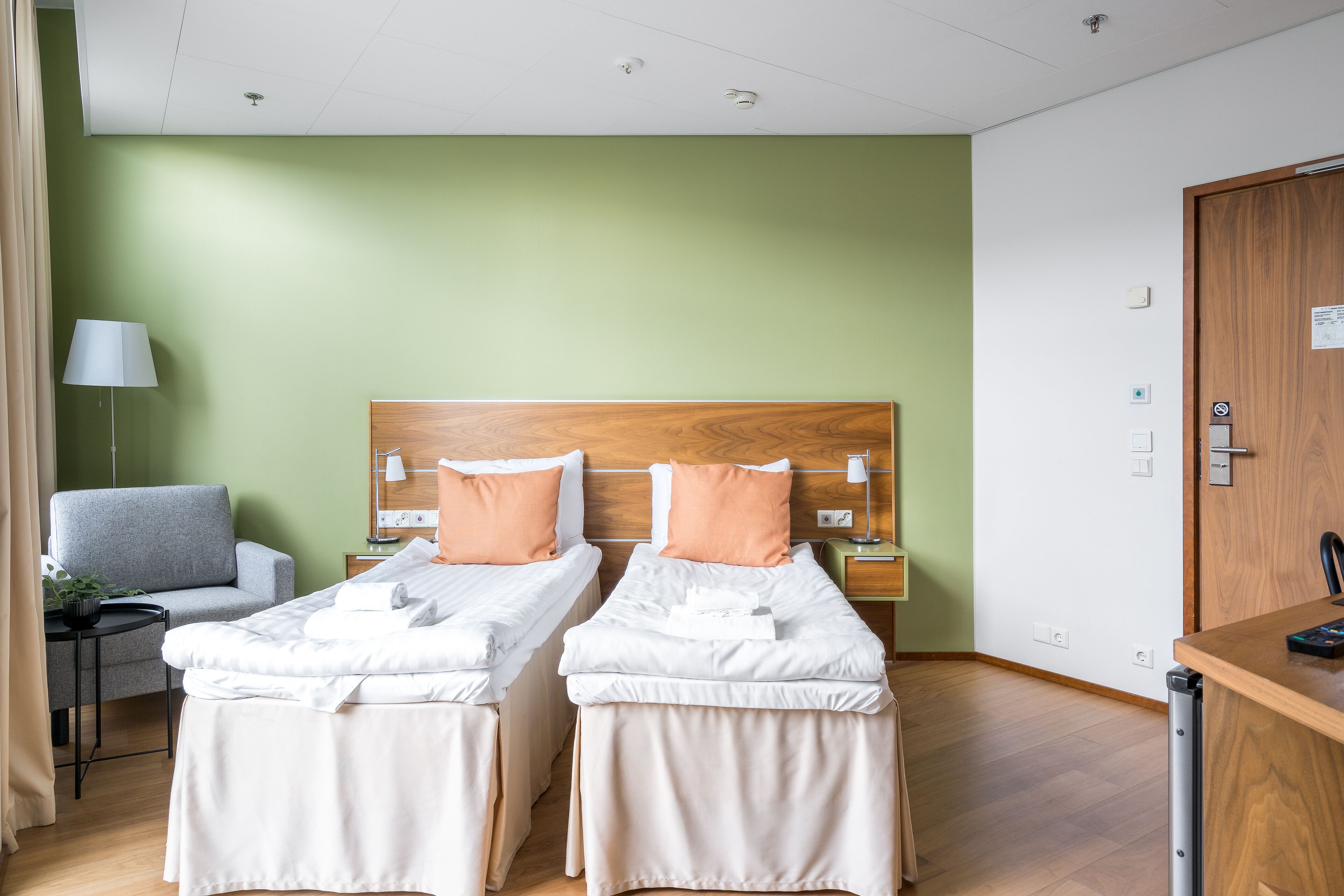 A hotel room with two single beds, a desk, a chair, and a lamp.
