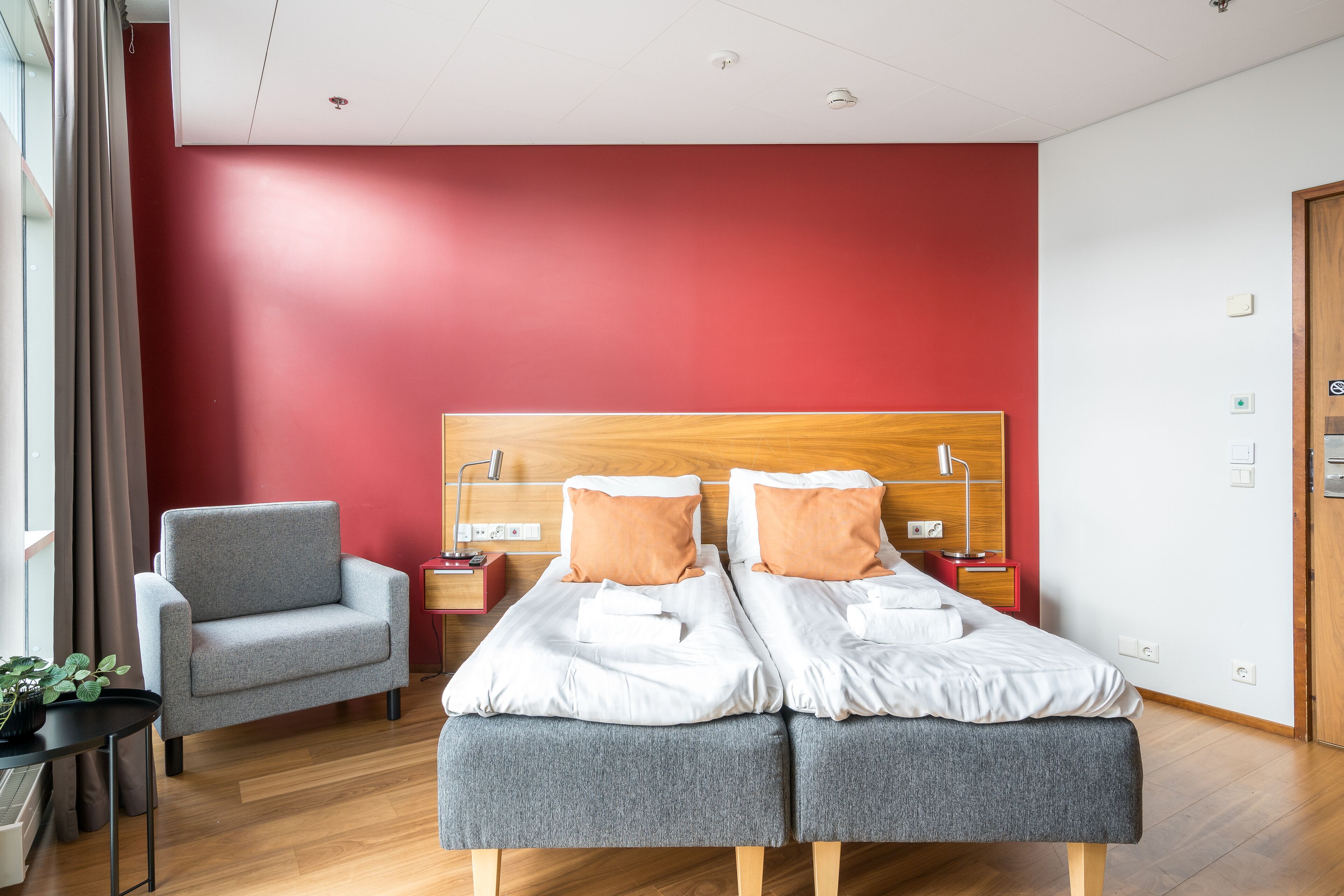 A modern hotel room with a red accent wall, a grey sofa, a wooden headboard, and a bedside table with a lamp.