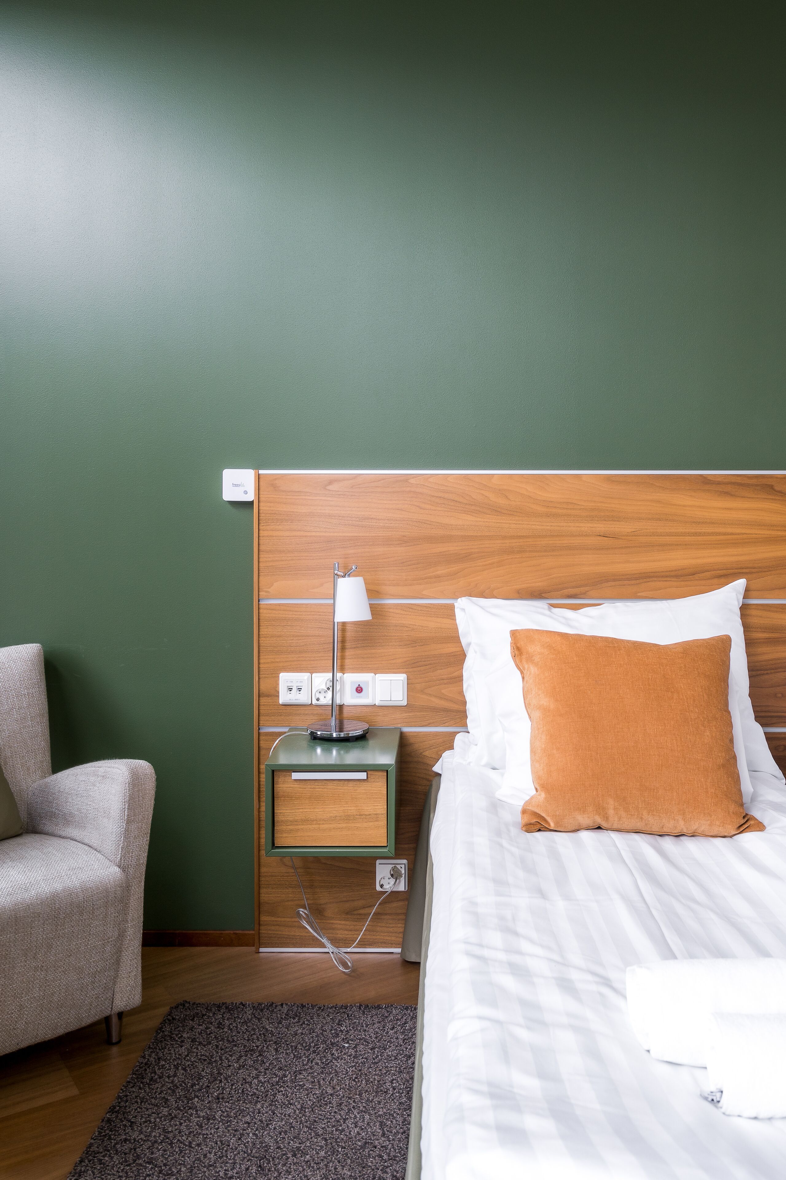 Una habitación de hotel con cabecera de madera, una cama blanca con almohada marrón, una mesita de noche con lámpara y una silla.