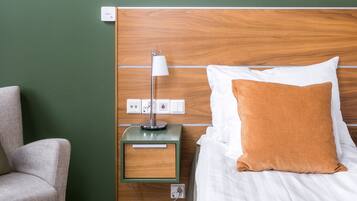 A hotel room with a wooden headboard, a white bed with a brown pillow, a bedside table with a lamp, and a chair.