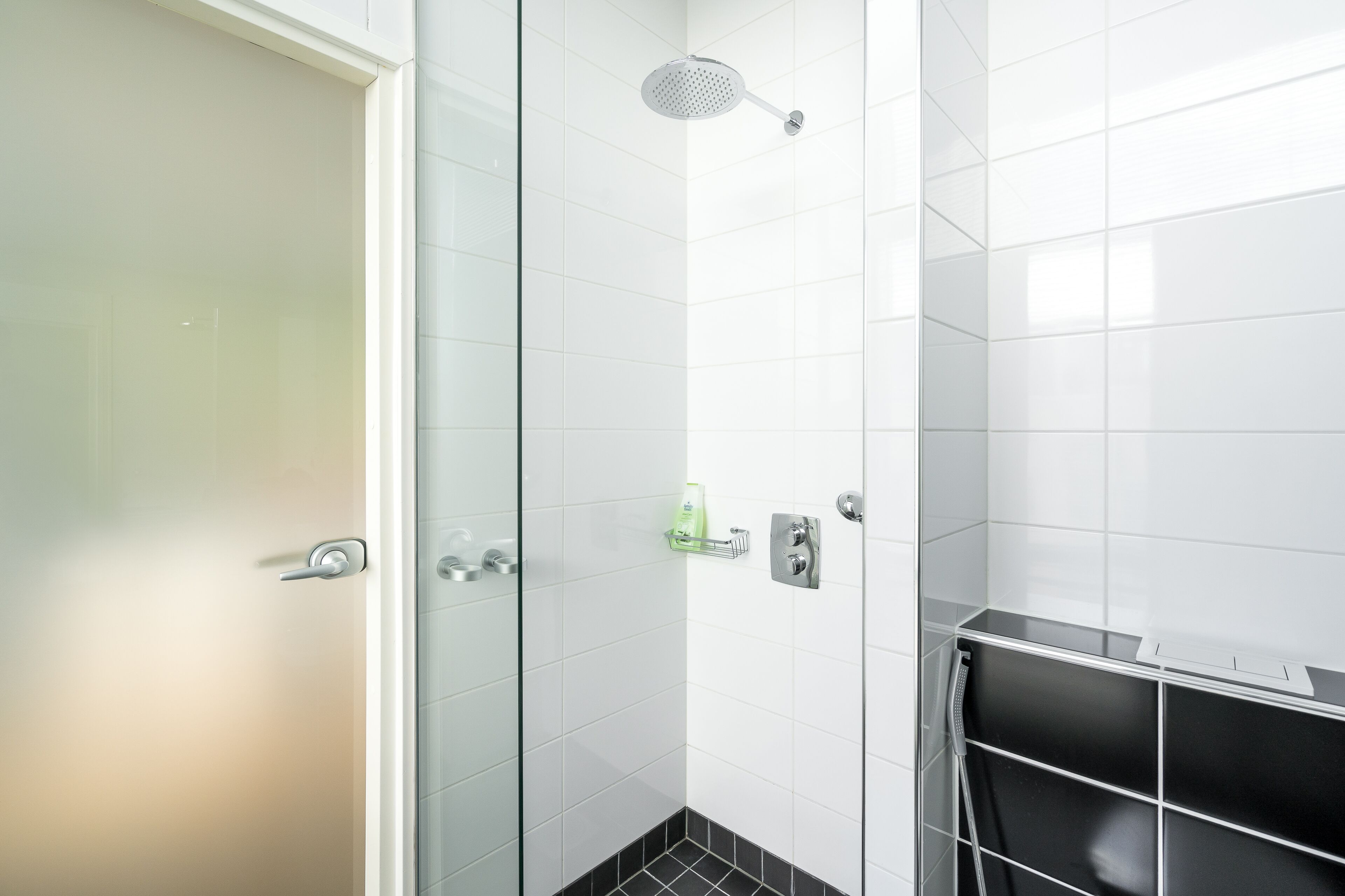 A modern bathroom with white tiles, a shower with a handheld showerhead, a glass shower door, and a black tiled floor.