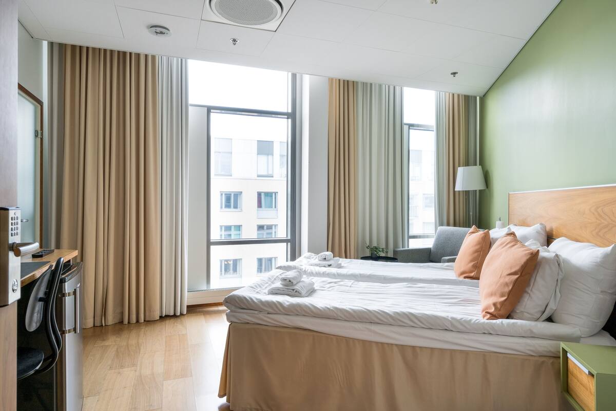 Ein Hotelzimmer mit Bett, Schreibtisch, Stuhl, Lampe und Blick auf die Stadt durch große Fenster.