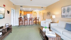A modern living room with a white sofa, a coffee table, and a dining area with wooden chairs and a kitchen in the background. - 3BR Golf View w/ $50 Theme Park Credit (Kissimmee)