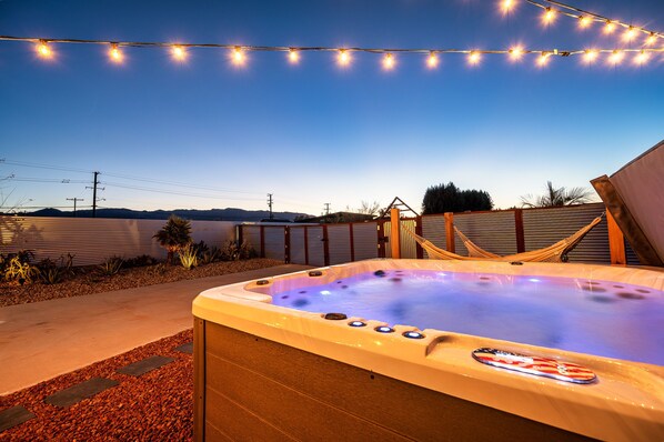 A hot tub with a logo, surrounded by a wooden deck, string lights, and a hammock.