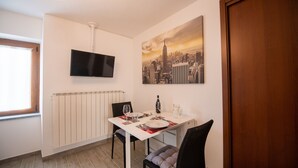 A dining area with a white table, black chairs, a TV mounted on the wall, and a cityscape painting.