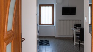 A narrow hallway leading to a kitchen with a window, a radiator, and a television on the wall.