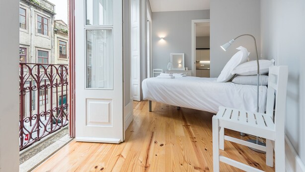 Une chambre à coucher avec un lit, une table de chevet, une lampe et une vue sur la rue, visible par la porte.