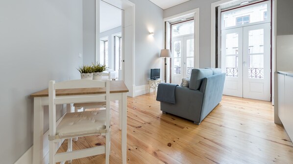 Un espace de vie moderne avec un plancher en bois, un canapé gris, une table à manger blanche avec des chaises et une petite télévision posée sur une table d’appoint.