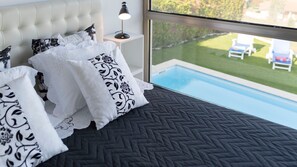 A bedroom with a view of a swimming pool, a bed with patterned pillows, and a bedside lamp.