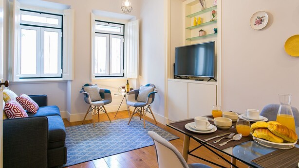 A modern living room with a sofa, two armchairs, a coffee table, and a TV. There are windows, a bookshelf, and a dining area with a table set for breakfast.