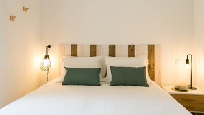 A neatly made bed with white bedding and two green pillows, a wooden headboard, a bedside table with a lamp, and a wall-mounted light fixture.