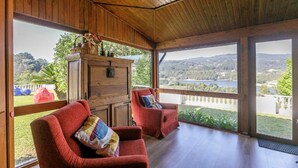 Amplia habitación con techo de madera y ventanas grandes que ofrecen vista a un lago y colinas. Hay dos sillones rojos y un aparador de madera con objetos decorativos.
