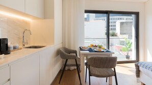 Una camera d'albergo compatta con angolo cottura, un'area pranzo con due sedie, un tavolo apparecchiato per la colazione e un balcone con vista sugli edifici.
