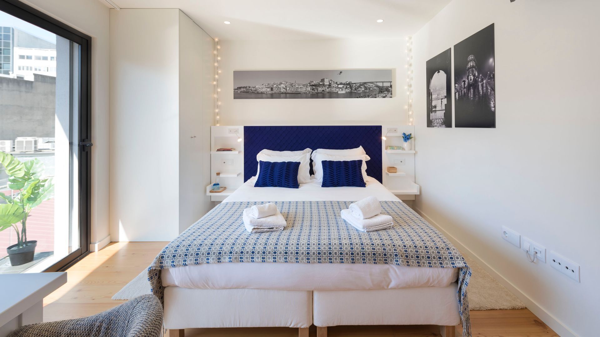 A modern hotel room with a large bed, a view of the city, and a cityscape painting on the wall.