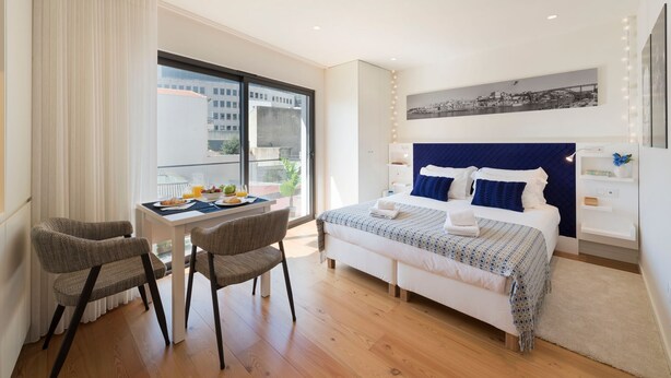 Una moderna camera d'hotel con un letto grande, un'area pranzo con tavolo e sedie, e vista sulla città dalla finestra.