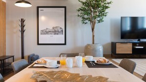 A modern dining area with a table set for breakfast, including croissants, coffee, and orange juice. A large vase and a framed picture are on the wall, and a TV is visible in the background. - Modern Loft w/ AC in Downtown Porto by LovelyStay (Porto)