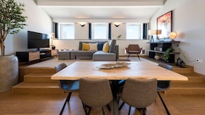 A modern living room with a large wooden dining table, grey sofas, and a flat-screen TV.
