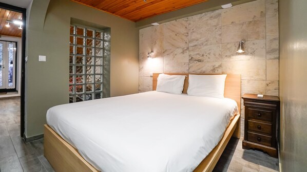 A bedroom with a large bed, wooden headboard, marble wall, and a small wooden nightstand with drawers.