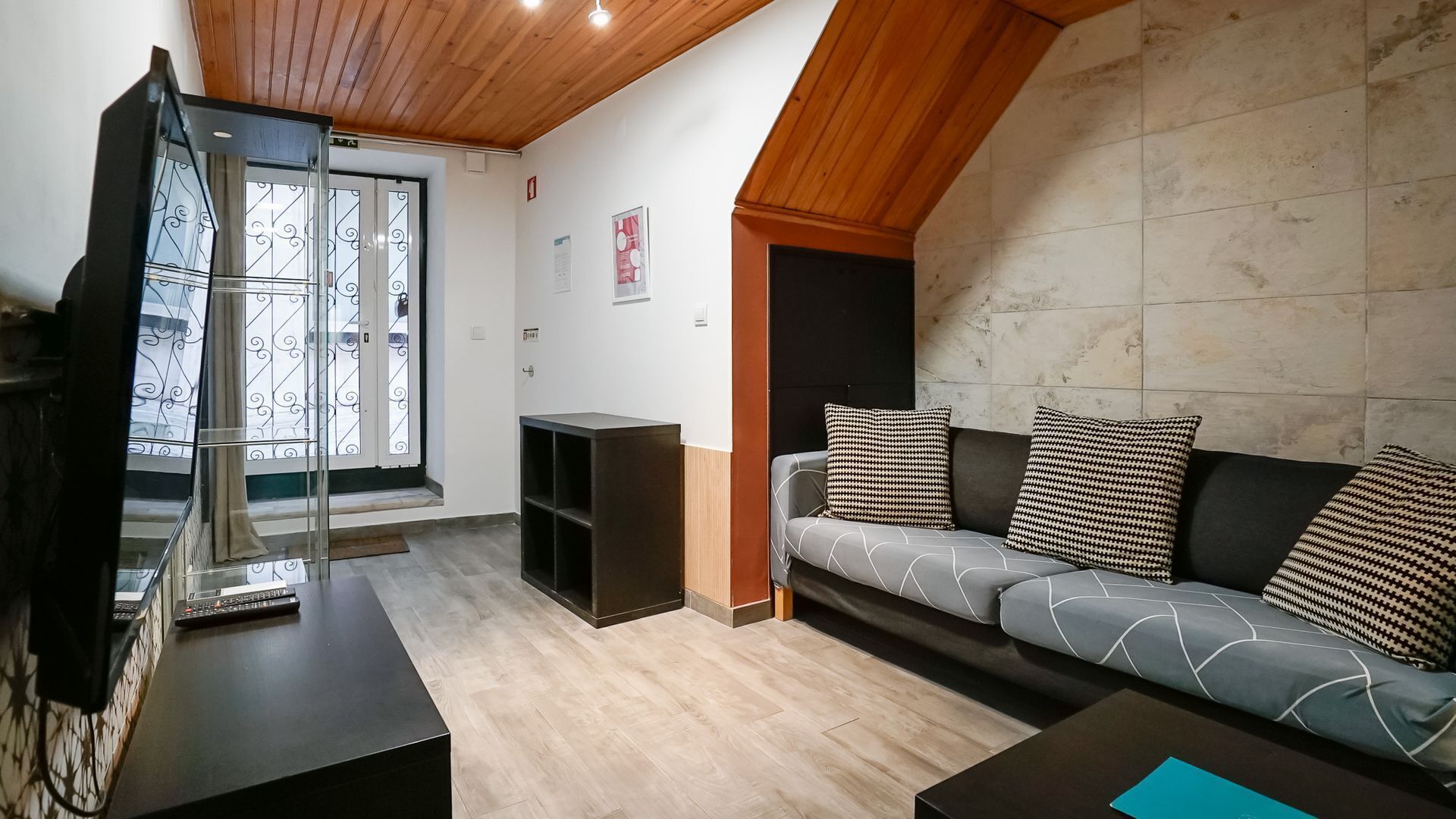 A modern living room with a wooden ceiling, a grey sofa with patterned cushions, a black entertainment unit, and a large window with decorative glass.