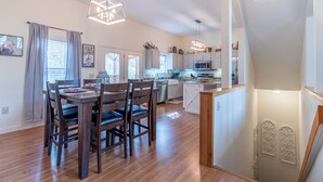 Una cocina moderna con armarios blancos, un comedor con mesa y sillas de madera, y lámparas colgantes.