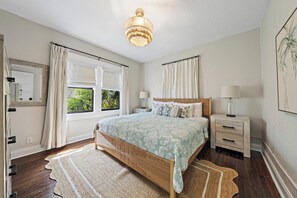 Un dormitorio con cama, mesitas de noche, lámpara de noche, candelabro y ventana con cortinas.