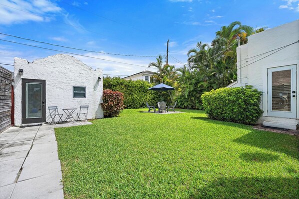 A well-maintained lawn with a patio table and chairs, surrounded by lush greenery and a white building with a door and window.