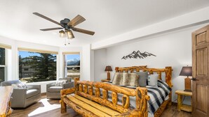 A bedroom with a wooden bed, a ceiling fan, and a view of mountains through large windows. - Spacious Family Getaway, King Beds, Hot Tub, RMNP (Estes Park)