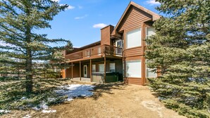 A two-story wooden cabin with a balcony, surrounded by evergreen trees and a snow-covered ground. - Spacious Family Getaway, King Beds, Hot Tub, RMNP (Estes Park)