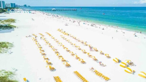 Een strand met rijen gele parasols en strandstoelen, een pier op de achtergrond en een helderblauwe lucht.