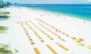 A beach with rows of yellow umbrellas and beach chairs, a pier in the background, and a clear blue sky.