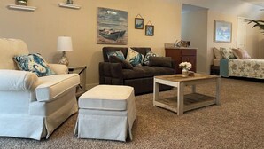 Een woonkamer met een beige bank, een donkerbruine fauteuil en een houten salontafel. Er zijn wandplanken met decoratieve objecten en een schilderij van een boot aan de muur.