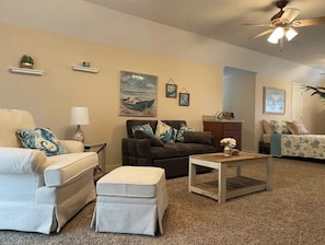 A living room with a beige sofa, a dark brown armchair, and a wooden coffee table. There are wall-mounted shelves with decorative items and a painting of a boat on the wall.