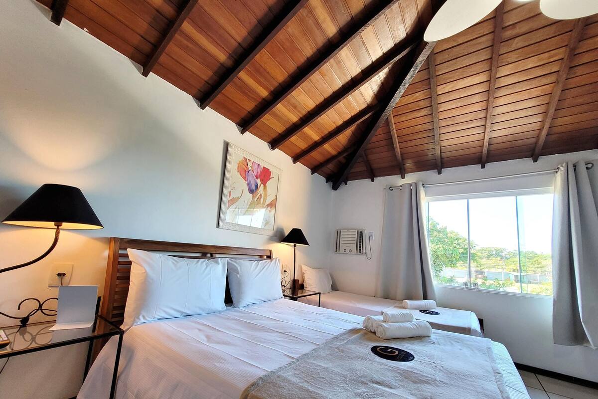 A hotel room with a wooden ceiling, a bed with white linens, a bedside table with a lamp, a window with curtains, and a painting on the wall.