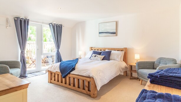 A bedroom with a wooden bed, two bedside tables, a chair, a sofa, and a view of the outdoors through a glass door. - The Croft (Thetford)