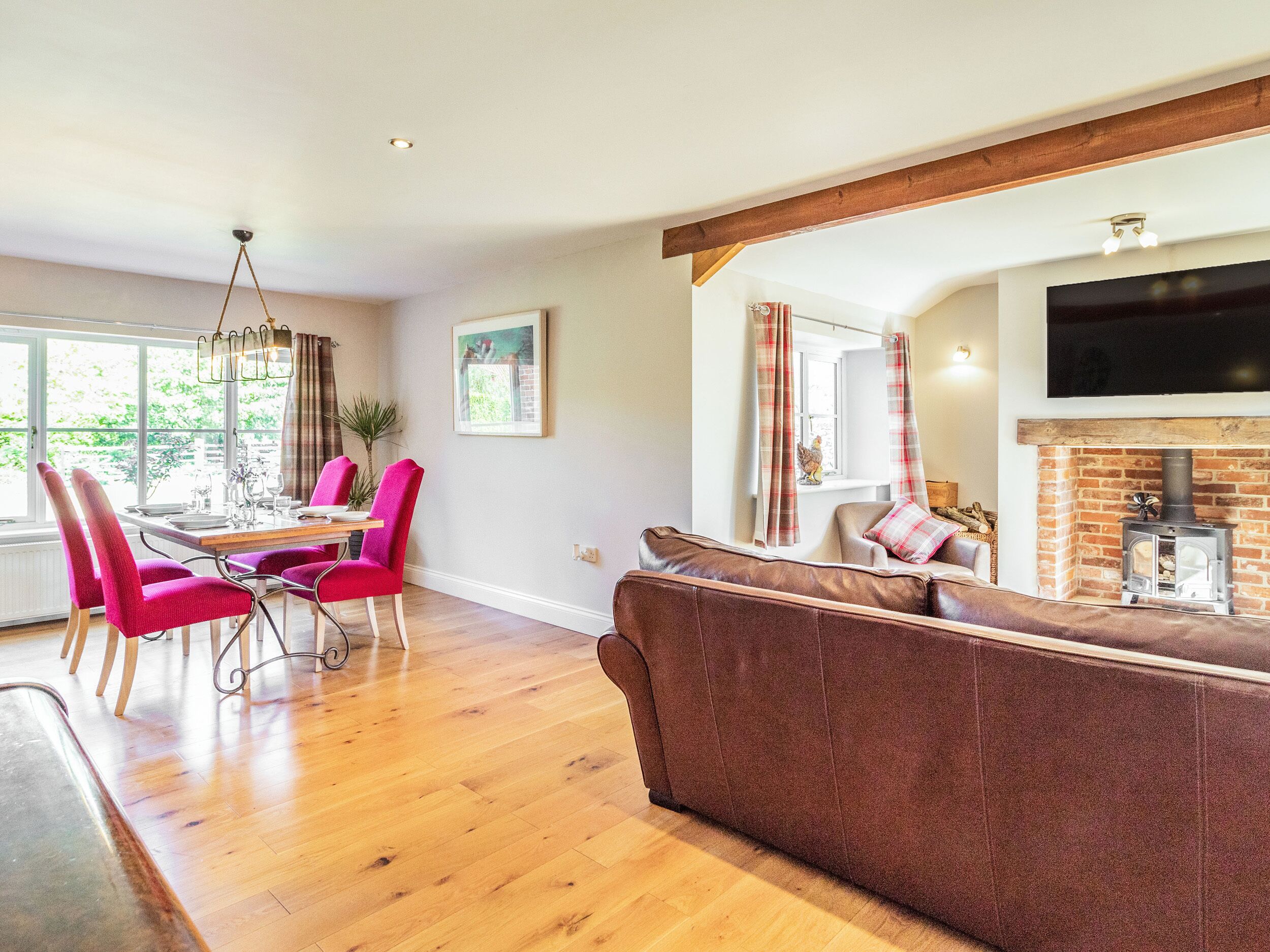 A modern living room with a wooden floor, a brown sofa, a dining area with pink chairs, and a fireplace with a TV above it.