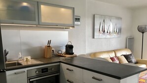 Une cuisine moderne équipée d’un four encastré, d’une machine à café et d’un évier. Le salon adjacent est doté d’un canapé beige agrémenté de coussins colorés.