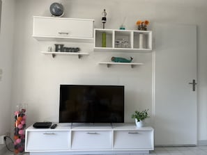 Una sala de estar moderna con un televisor de pantalla plana, muebles blancos y objetos decorativos en estantes.