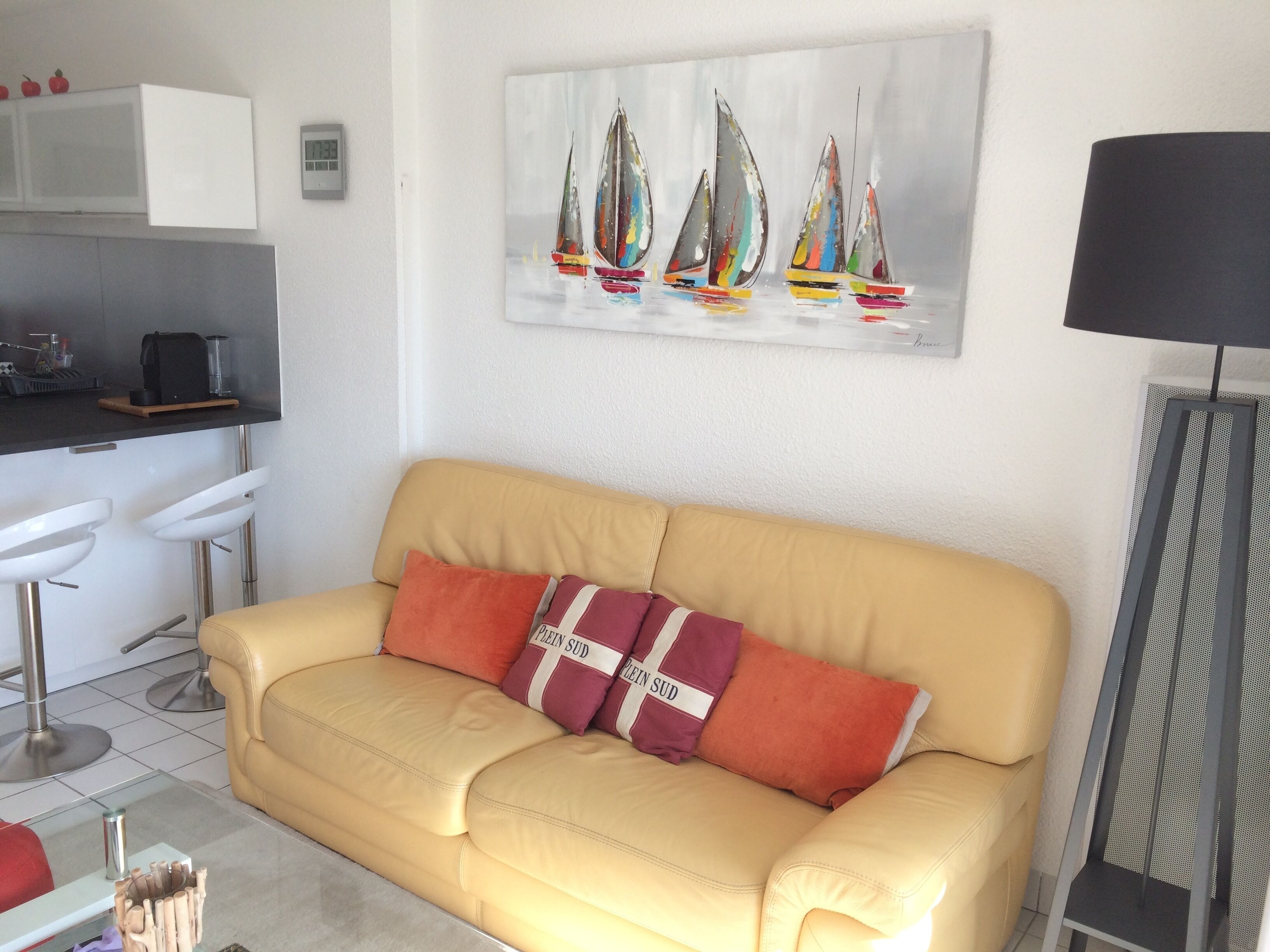 A living room with a yellow sofa, two red cushions, a painting of sailboats, a kitchen area with white cabinets, and a black floor lamp.