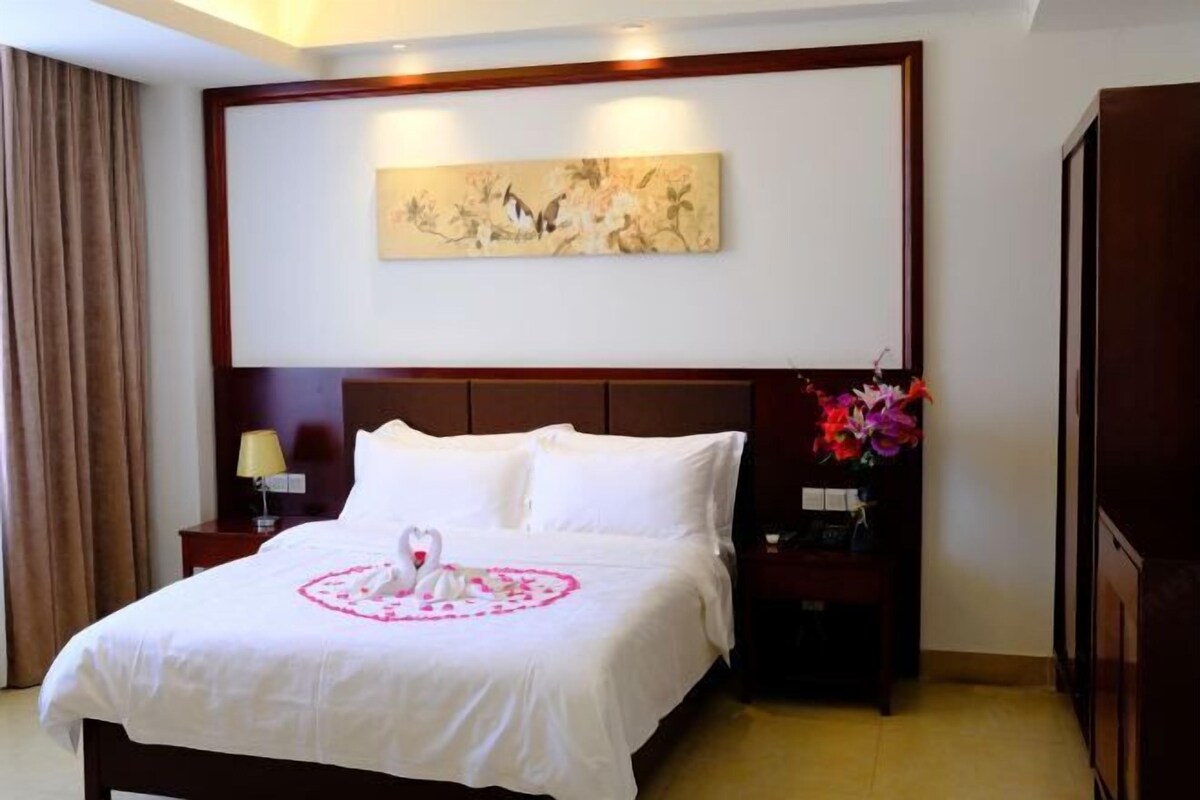 A hotel room with a bed, a nightstand, a lamp, a painting on the wall, and a vase with flowers.