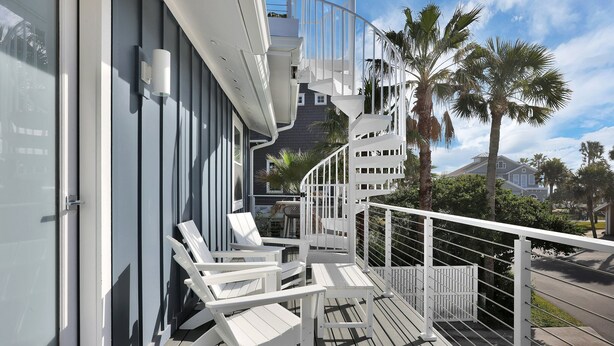 Un balcone con sedie e tavolo bianchi, una scala a chiocciola e vista su palme e una casa.