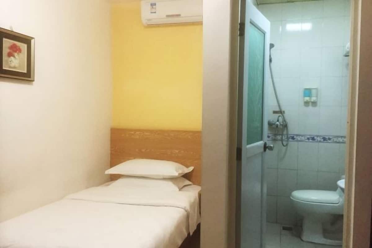 A small, single-bed room with a plain bed, a wall-mounted air conditioner, and a door leading to a bathroom with a toilet and shower.