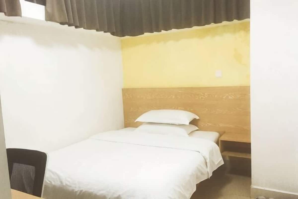 A small, single-bed room with a wooden headboard, a chair, and a window with curtains.