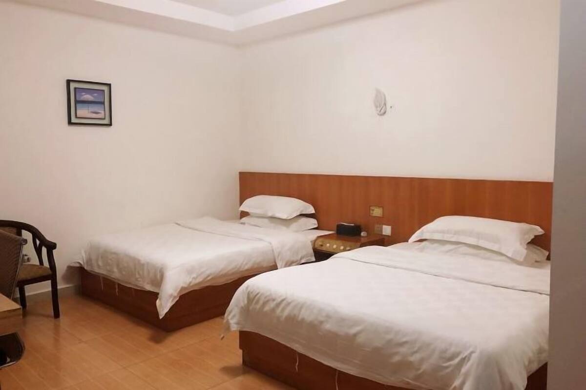 A hotel room with two single beds, a wooden headboard, a small table with a lamp, and a framed picture on the wall.
