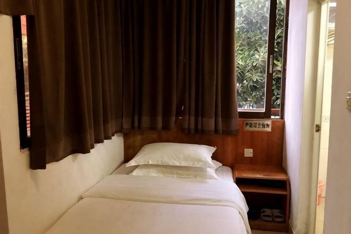 A single bed with a wooden headboard, a window with curtains, and a small shelf with a sign.