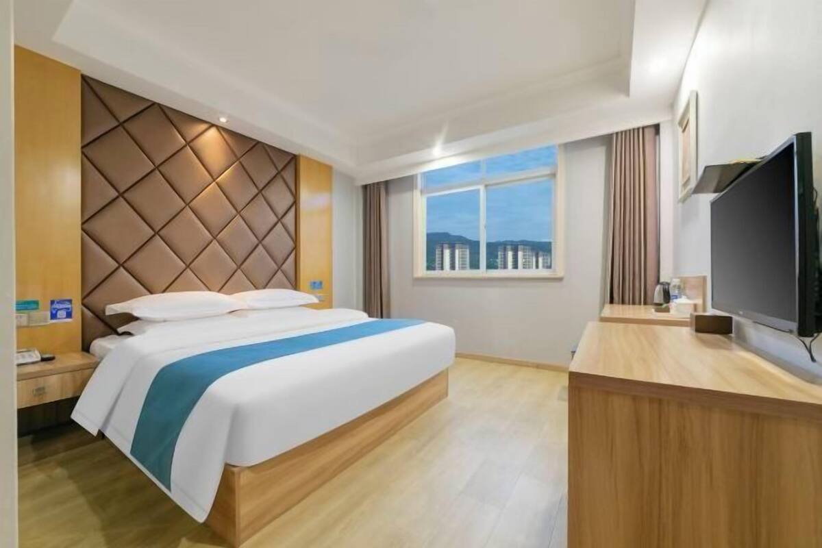 A hotel room with a bed, a wooden desk, a television, and a window with a view of buildings.