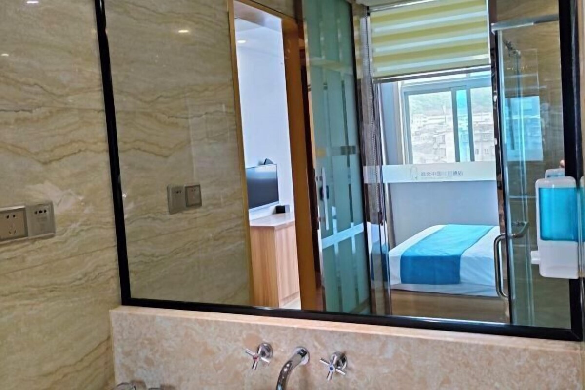 A bathroom with a marble wall, a glass shower enclosure, and a bed with a blue bedspread visible through the glass.